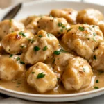 Plate Of Chicken And Herb Dumplings In Creamy Gravy Garnished With Fresh Parsley, Served On A White Plate With A Silver Spoon
