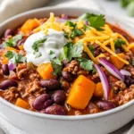 Bowl Of Ground Turkey Chili With Butternut Squash Topped With Shredded Cheese, Cilantro, Red Onion, And Sour Cream, On A Neutral Tabletop