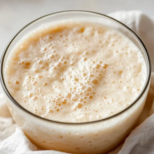 Bubbling How To Make Sourdough Starter In A Glass Bowl With A Kitchen Towel Beside It, Highlighting Its Airy Texture.