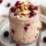 Jar Of Vanilla Cranberry Overnight Oats Layered With Yogurt And Topped With Fresh Berries, Almonds, And Cranberries On A White Plate