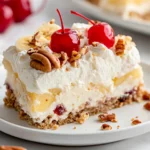 Slice Of Banana Split Cake Dessert On A White Plate, Topped With Whipped Cream, Pecans, And Maraschino Cherries